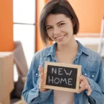 אישה מחזיקה שלט NEW HOME בין ארגזי מעבר דירה
