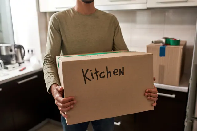 גבר נושא ארגז קרטון מסומן Kitchen במטבח ביום הובלת מקרר