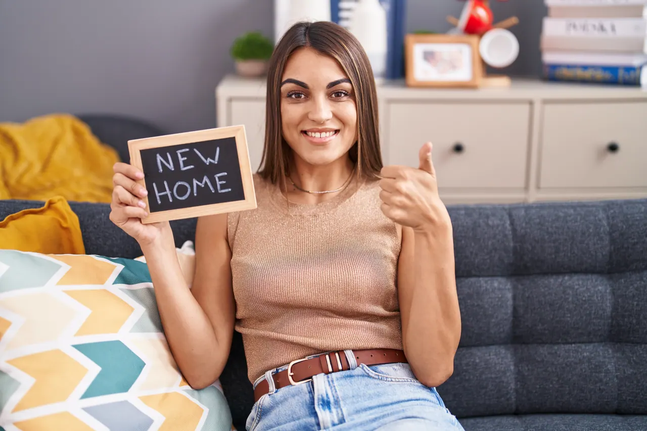 אישה יושבת על ספה ומחזיקה לוח גיר עם הכיתוב NEW HOME