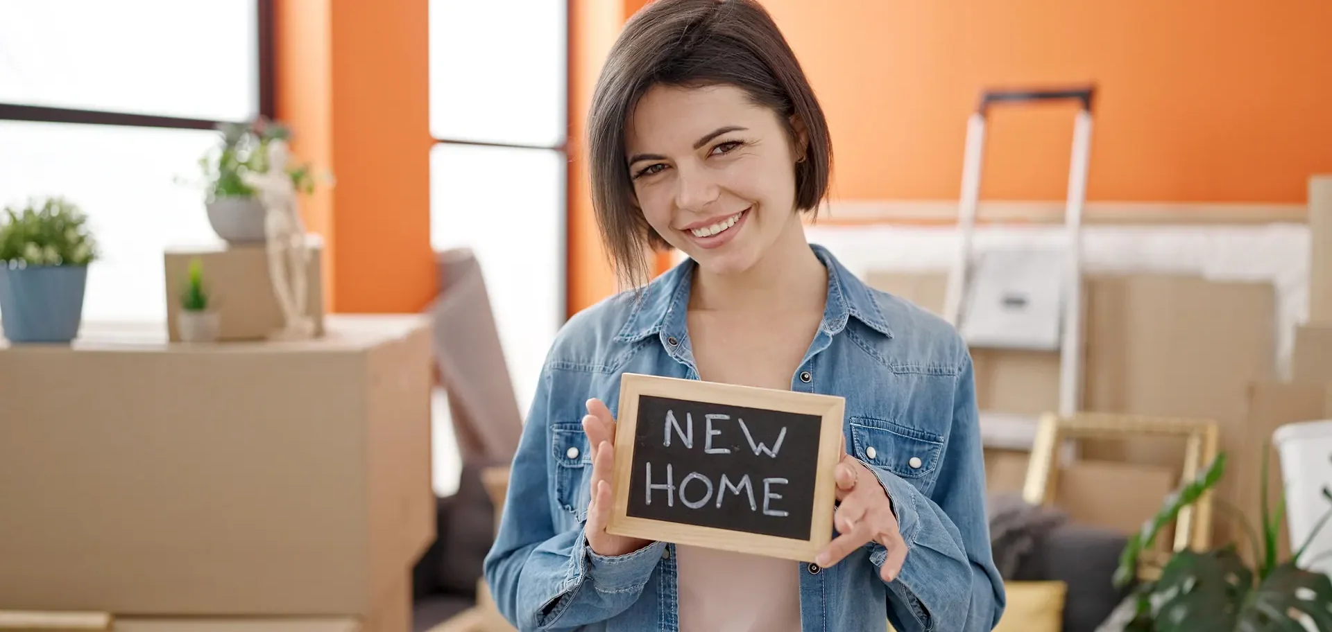 אישה מחזיקה שלט NEW HOME בדירה חדשה בין ארגזי מעבר