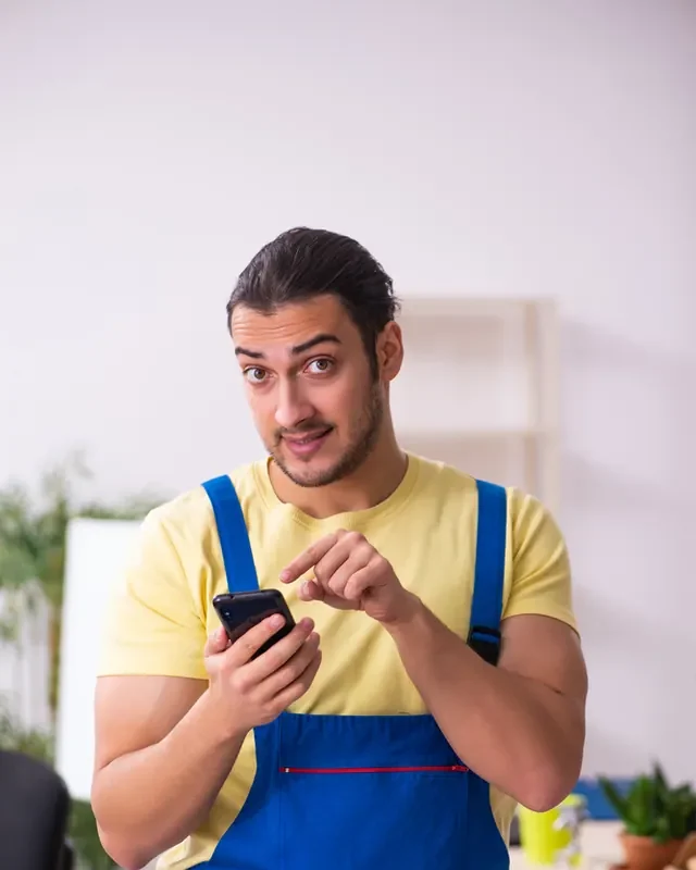 מוביל בסרבל עבודה כחול וחולצה צהובה מקליד בטלפון נייד
