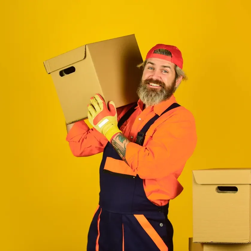 מוביל בסרבל עבודה וכפפות עבודה אוחז ארגז קרטון על רקע צהוב