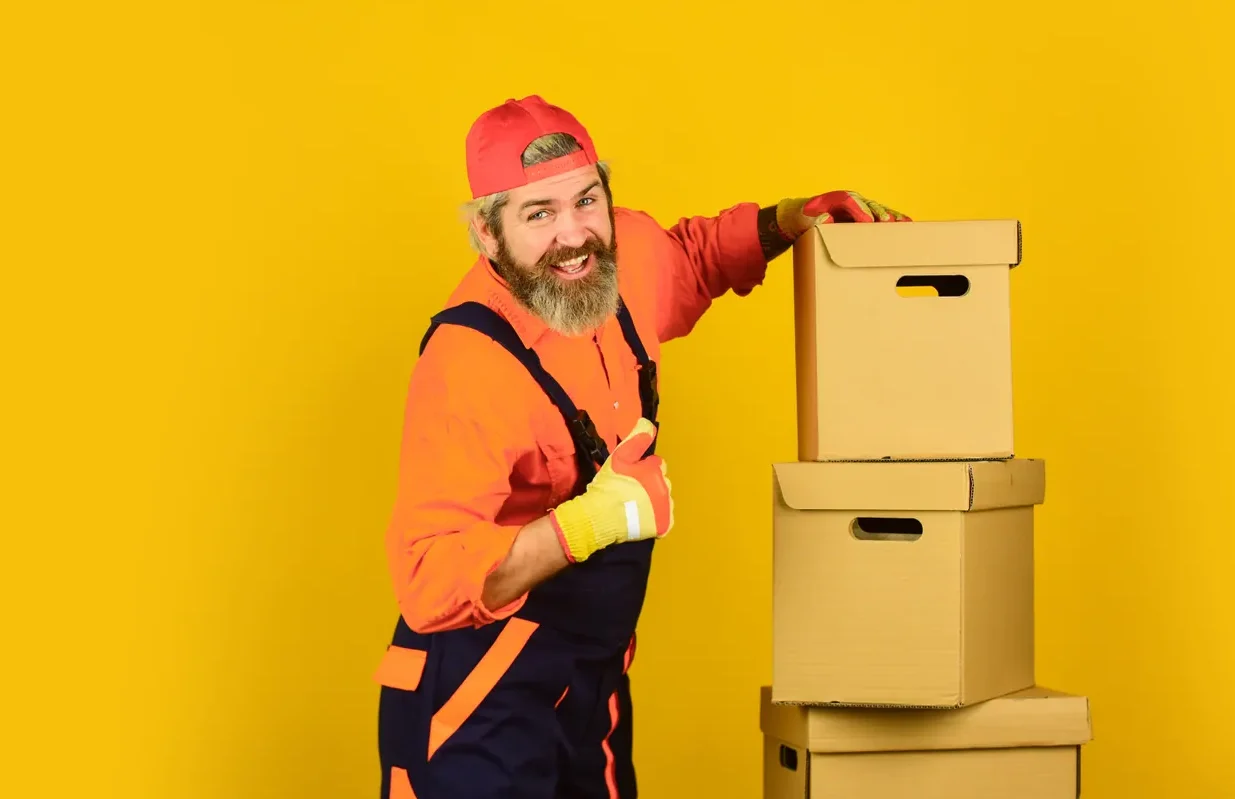 מוביל בסרבל כתום מרים אגודל ליד ערימת ארגזי קרטון על רקע צהוב
