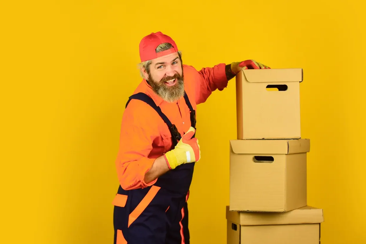 מוביל בסרבל כתום מרים אגודל ונשען על ארגזי קרטון מוערמים