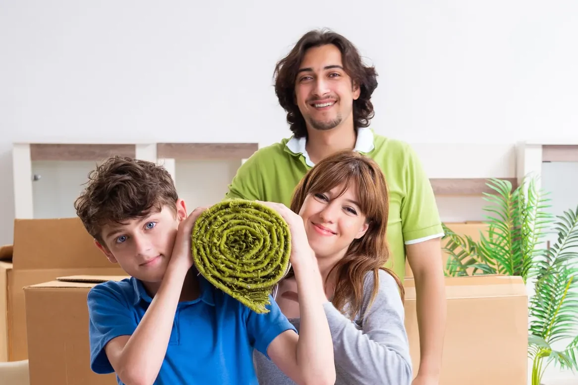 שלושה אנשים נושאים על הכתף שטיח ירוק מגולגל בחדר עם ארגזים