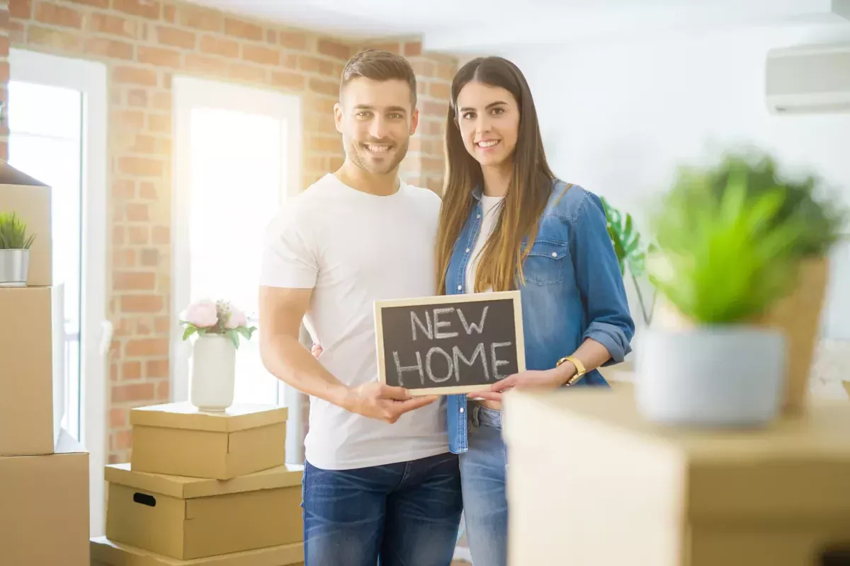 גבר ואישה מחזיקים לוח עם הכיתוב NEW HOME בין ארגזי קרטון בדירה