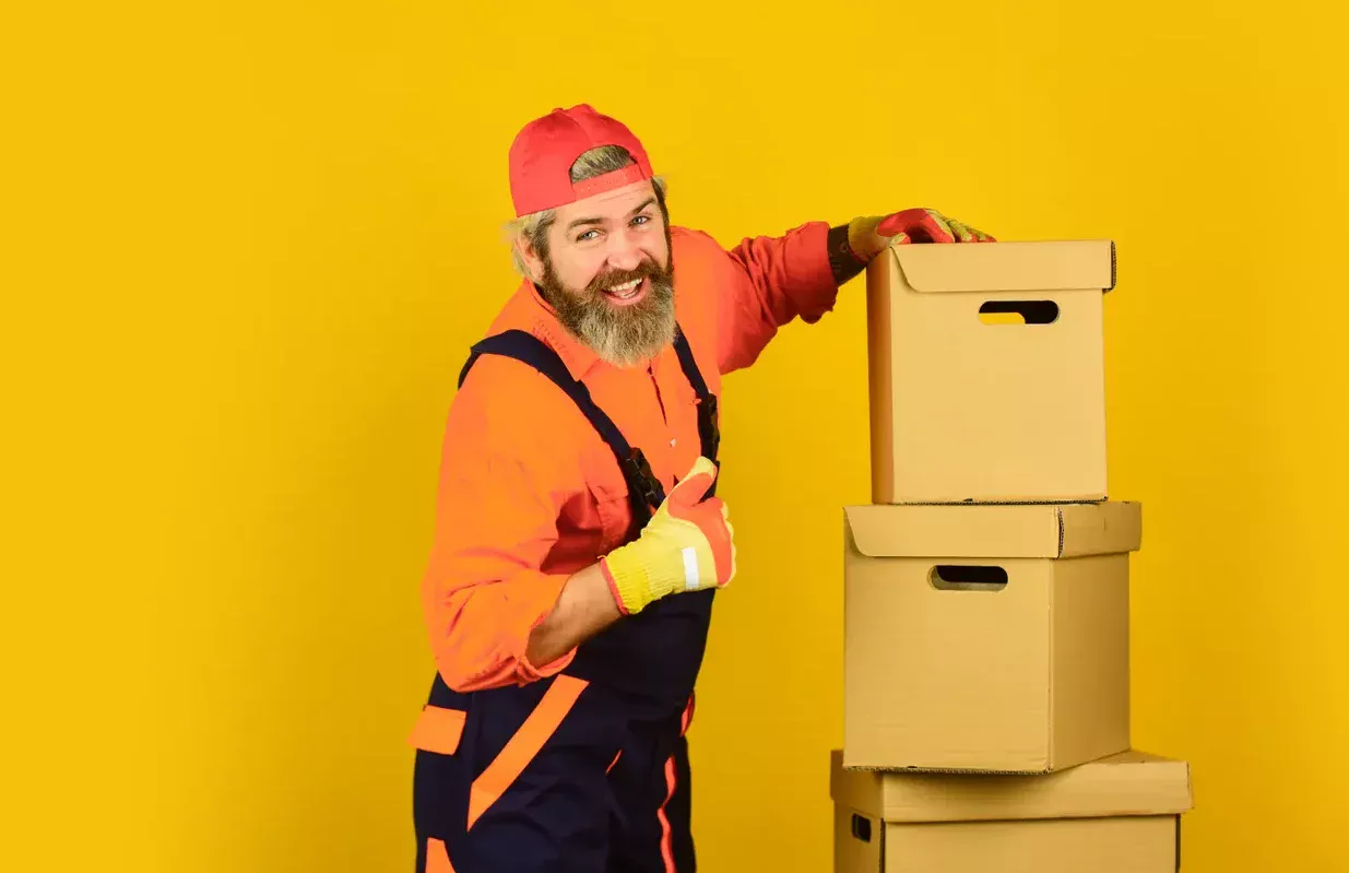 מוביל בסרבל כתום מרים אגודל ליד ערימת ארגזי קרטון על רקע צהוב
